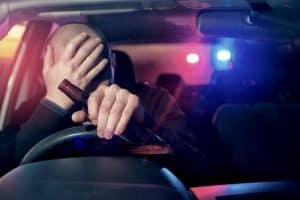 Man behind the wheel of a car holding a beer bottle while police car lights are seen in the background