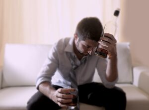 Upset man sitting on the bed holding a bottle of alcohol in his hand and a glass in the other